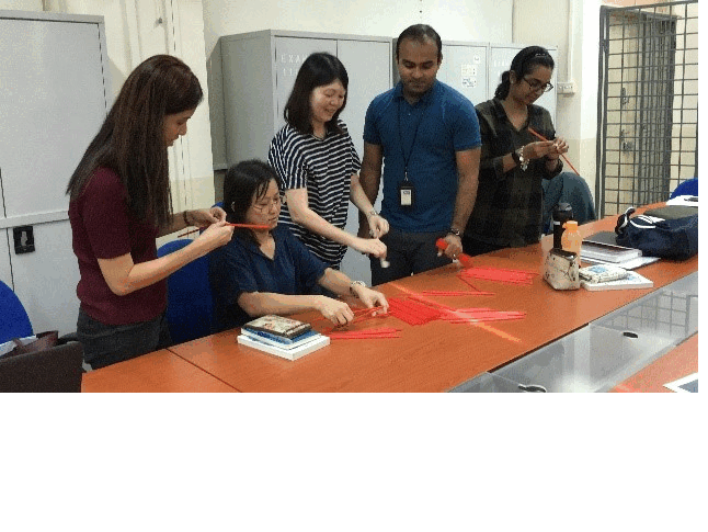 Five people gathered around an orange table handling red paper strips in an indoor room with lockers.
